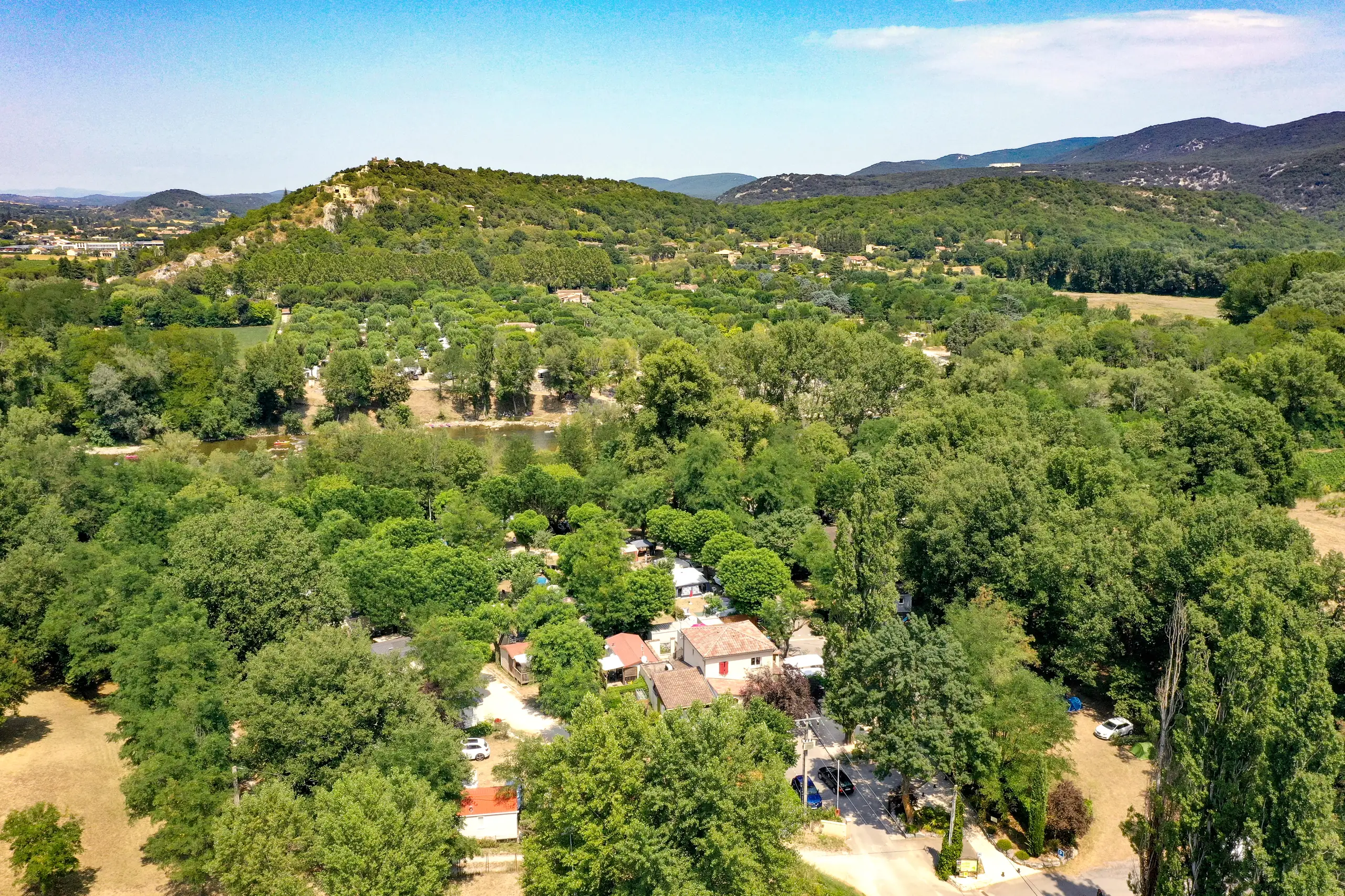 Le camping vu du ciel près de Vallon-Pont-d'Arc, ses grands arbres, l'Ardèche derrière les feuillages et les collines au loin