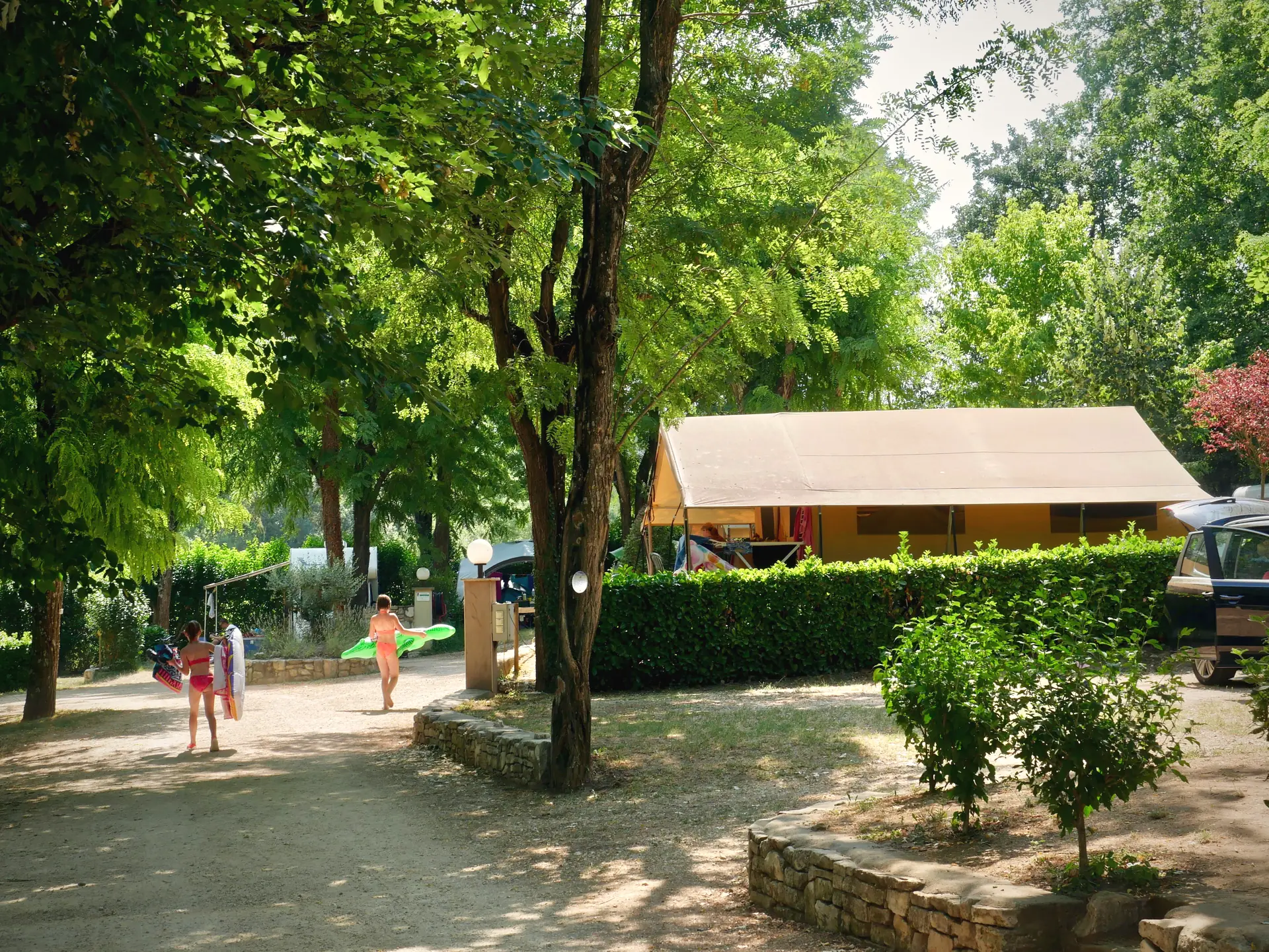 Ein schattiger Weg auf dem Campingplatz, ein Lodgezelt hinter seiner Hecke