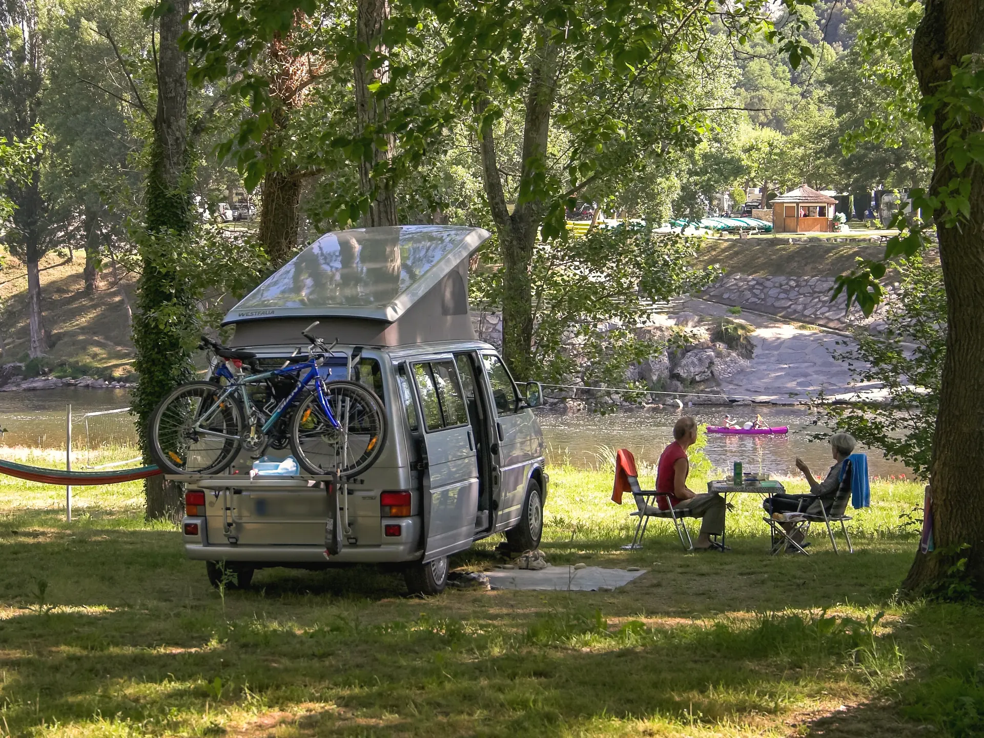 A campervan on a shaded pitch by the Ardèche, two holidaymakers at a table facing the river