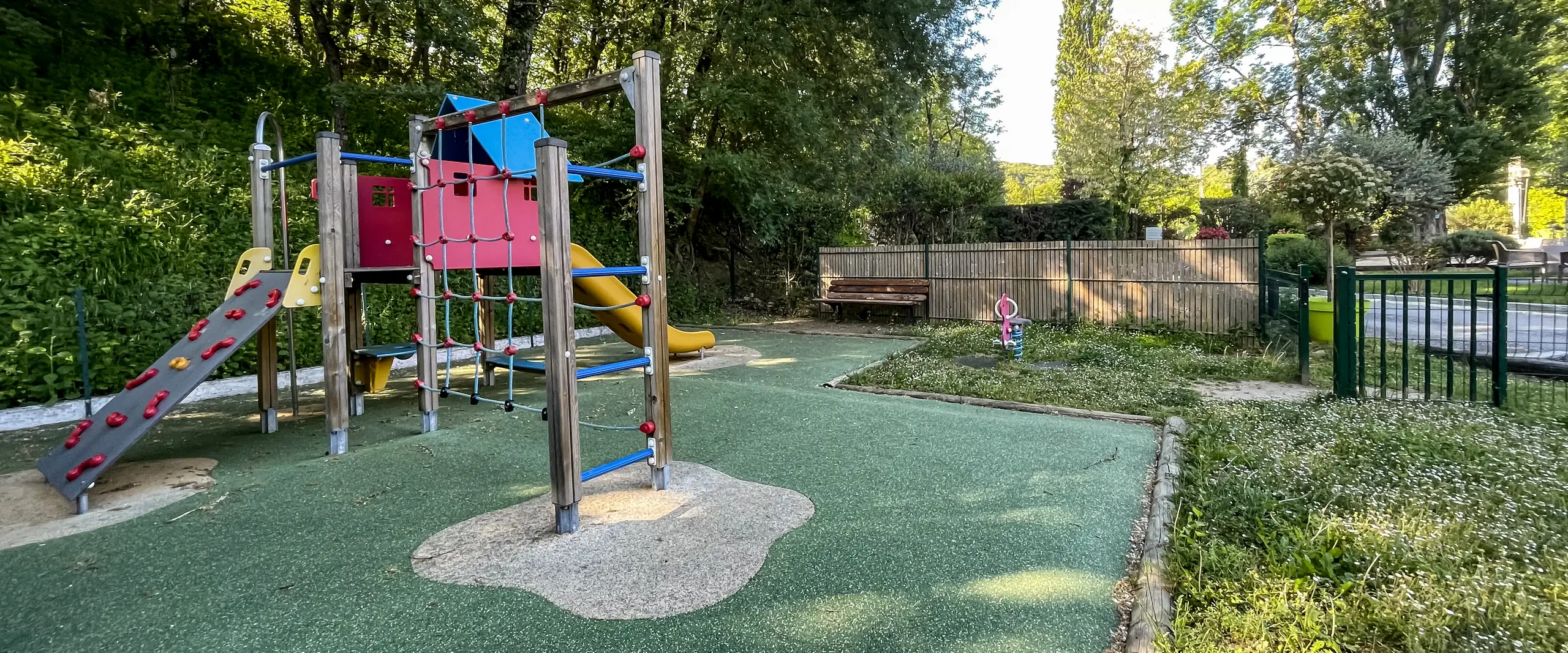 The campsite playground, its play frame with a slide and climbing ramp on green ground, in the shade of the trees