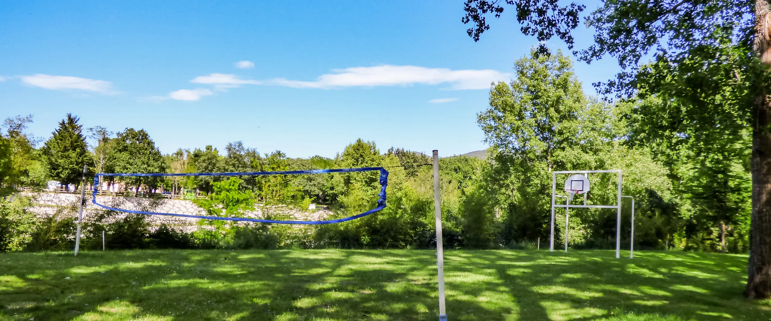 Terrain de sport en herbe du camping, avec filet de volley et panier de basket à l'ombre des arbres