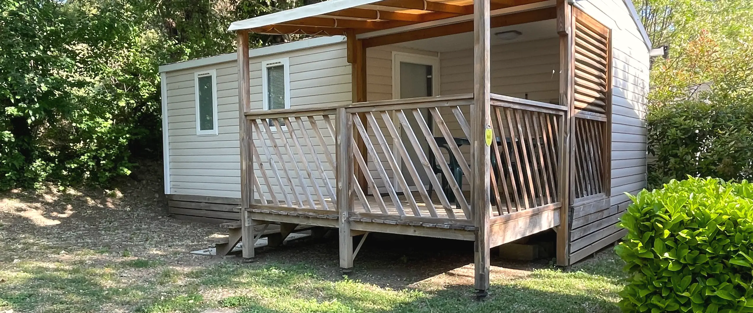A Classic mobile home with light cladding, a covered wooden terrace and a lattice railing, under the trees