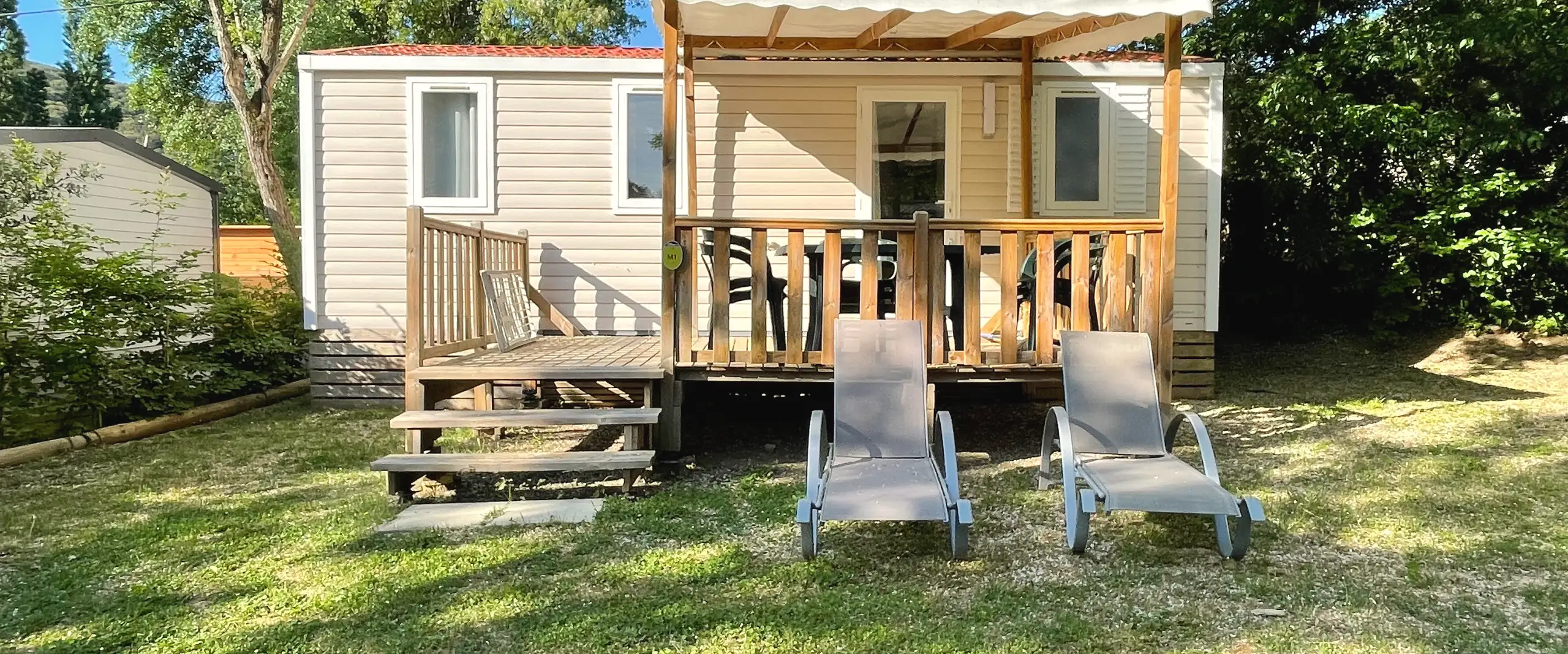 Mobil-home Classique en bardage clair, sa terrasse en bois sous une pergola, deux transats sur la pelouse