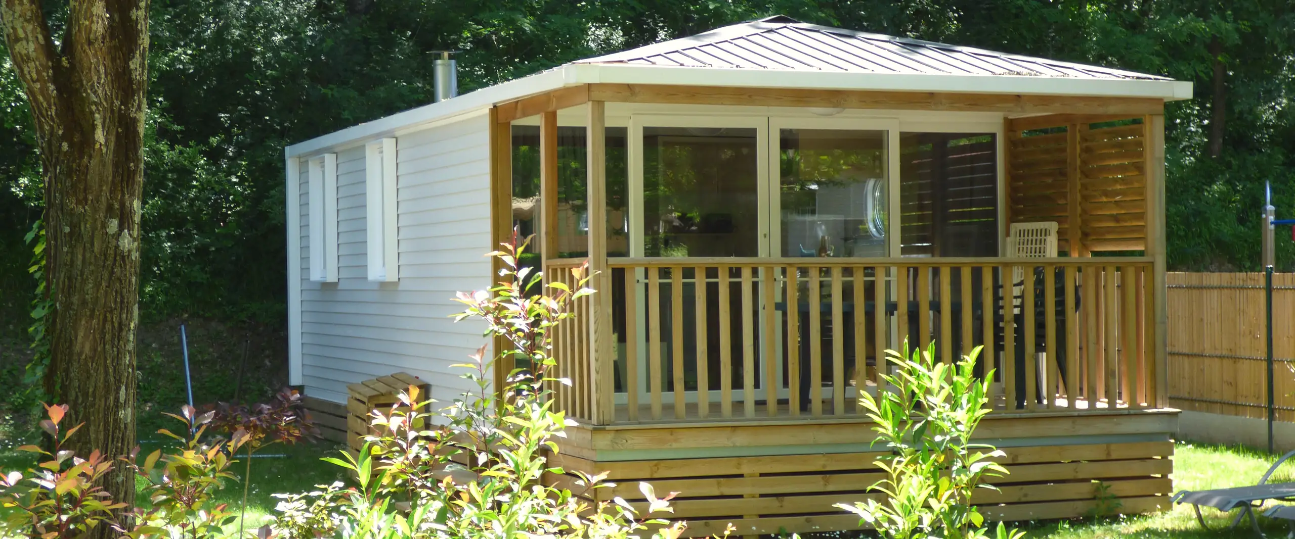 A Premium mobile home and its covered terrace with large windows, behind a flowering hedge, under the trees