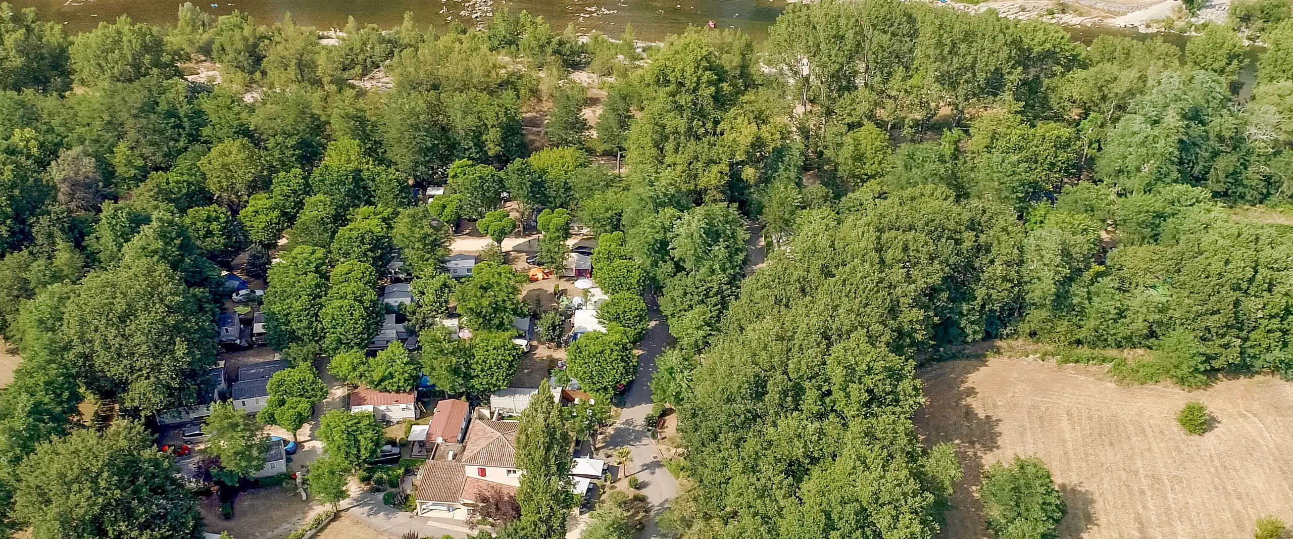 Le camping vu du ciel, ses allées et ses bâtiments sous les arbres, entre la rivière et la route