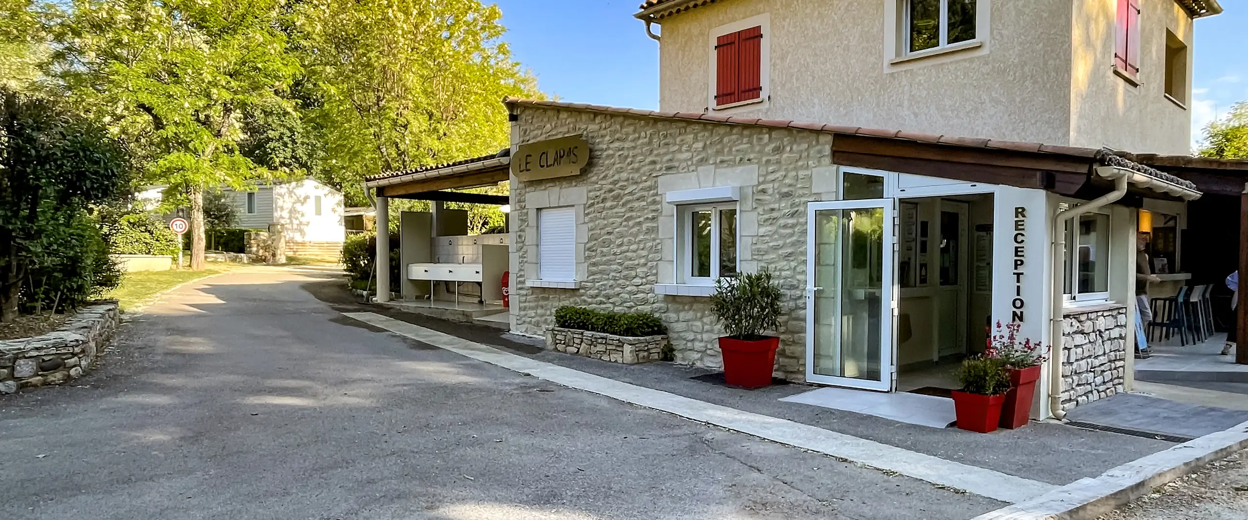 Bâtiment d'accueil du Camping Le Clapas en pierre, avec ses volets rouges et sa réception fleurie
