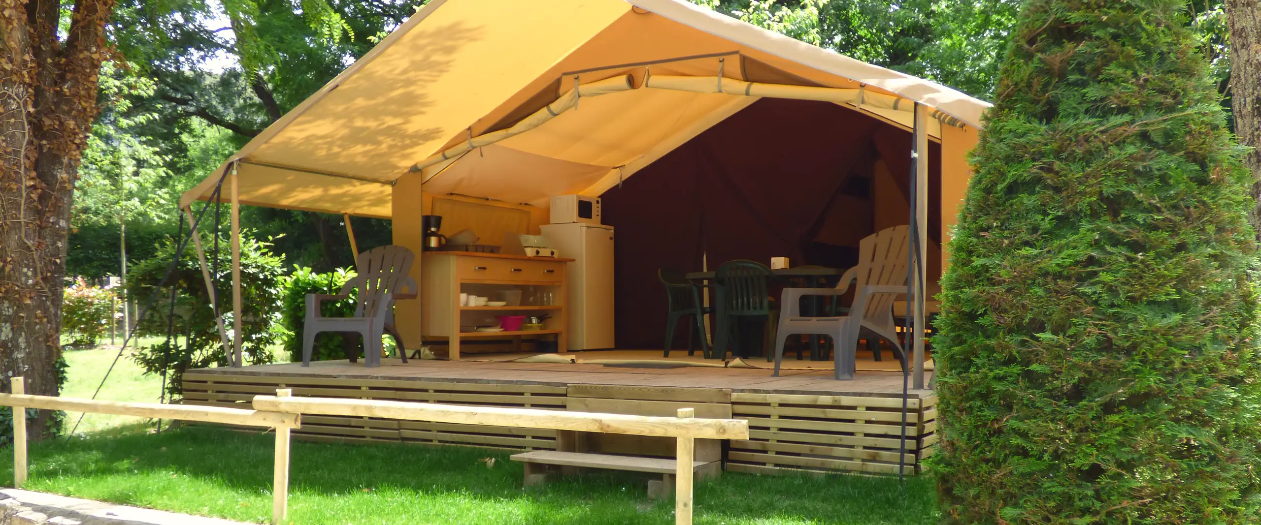 Tente Lodge en toile ouverte sur sa terrasse en bois couverte, avec coin cuisine, table et fauteuils derrière un muret en pierres