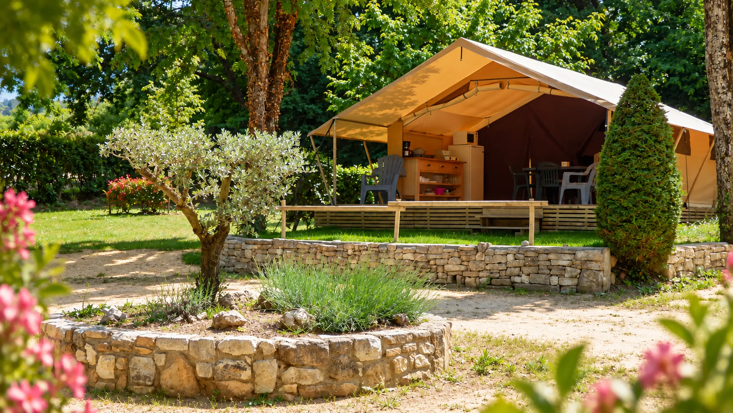 Ein Lodgezelt auf seiner Holzterrasse unter den Bäumen, hinter einem Beet mit Trockensteinmauer und einem Olivenbaum
