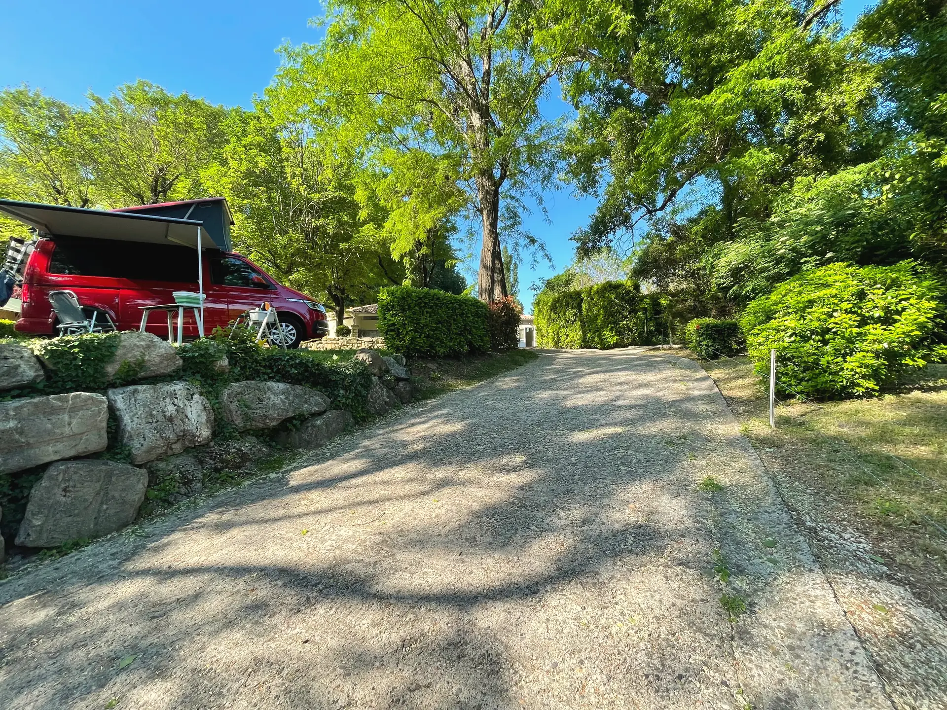 Allée gravillonnée du camping le long d'un muret de gros blocs de pierre, un van aménagé sur son emplacement
