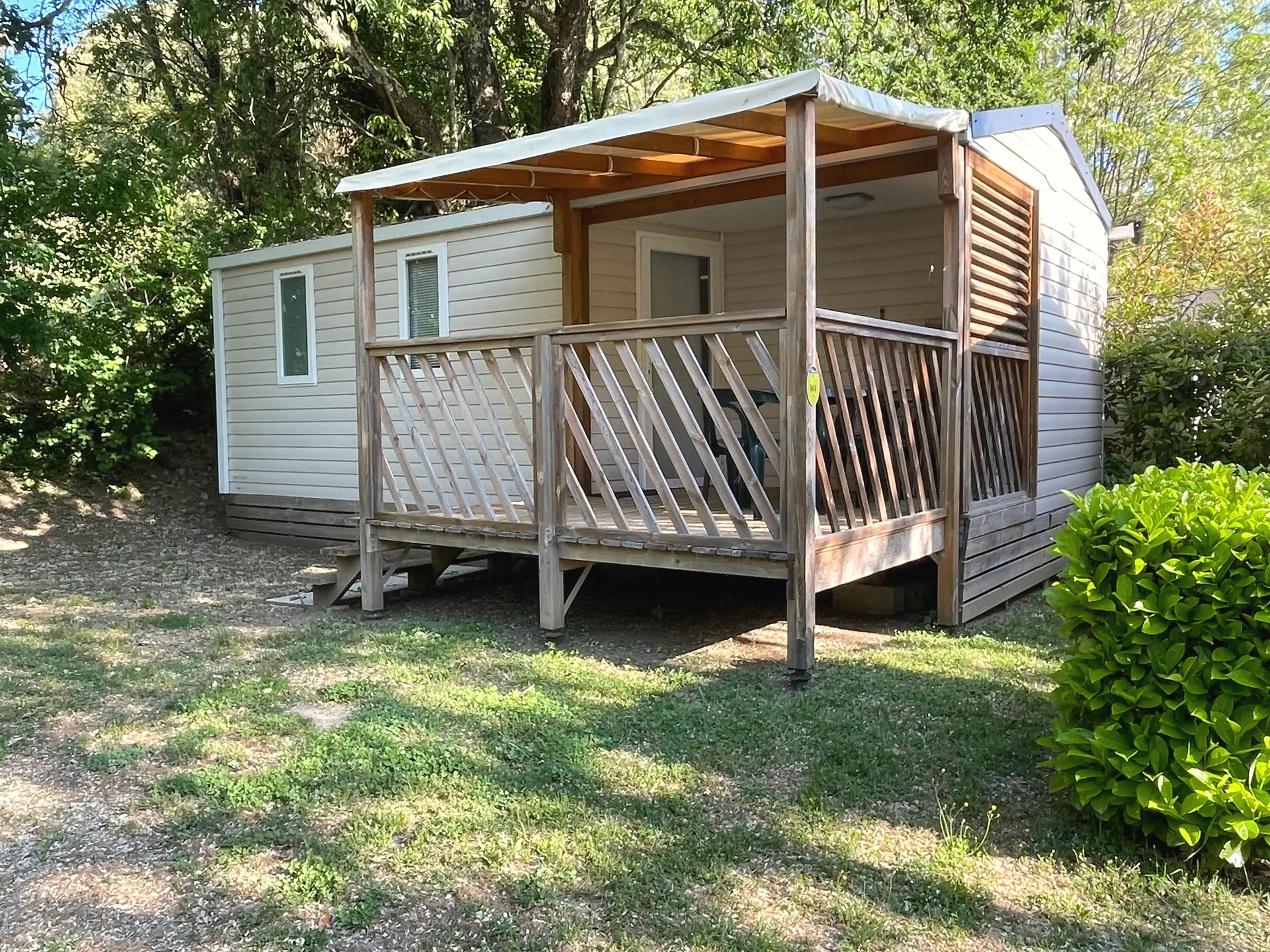 A Classic mobile home with light cladding, a covered corner terrace and a lattice railing, under the trees