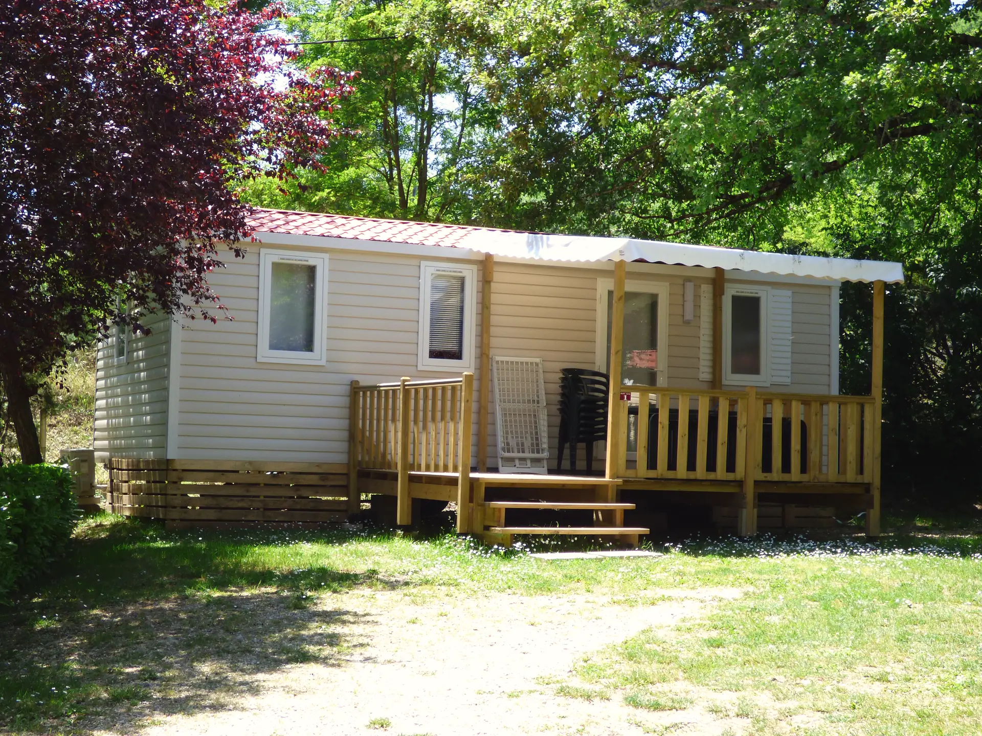 Mobil-home Classique vu de l'allée, sa terrasse en bois au milieu de la façade, sous les arbres