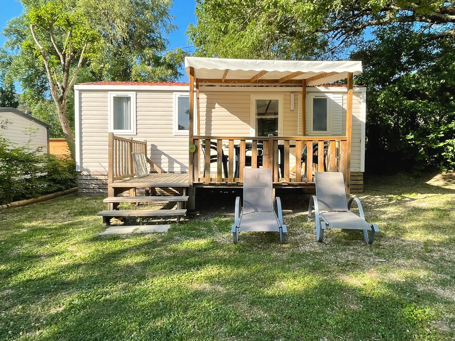 Mobil-home Classique en bardage clair, sa terrasse en bois sous une pergola, deux transats sur la pelouse