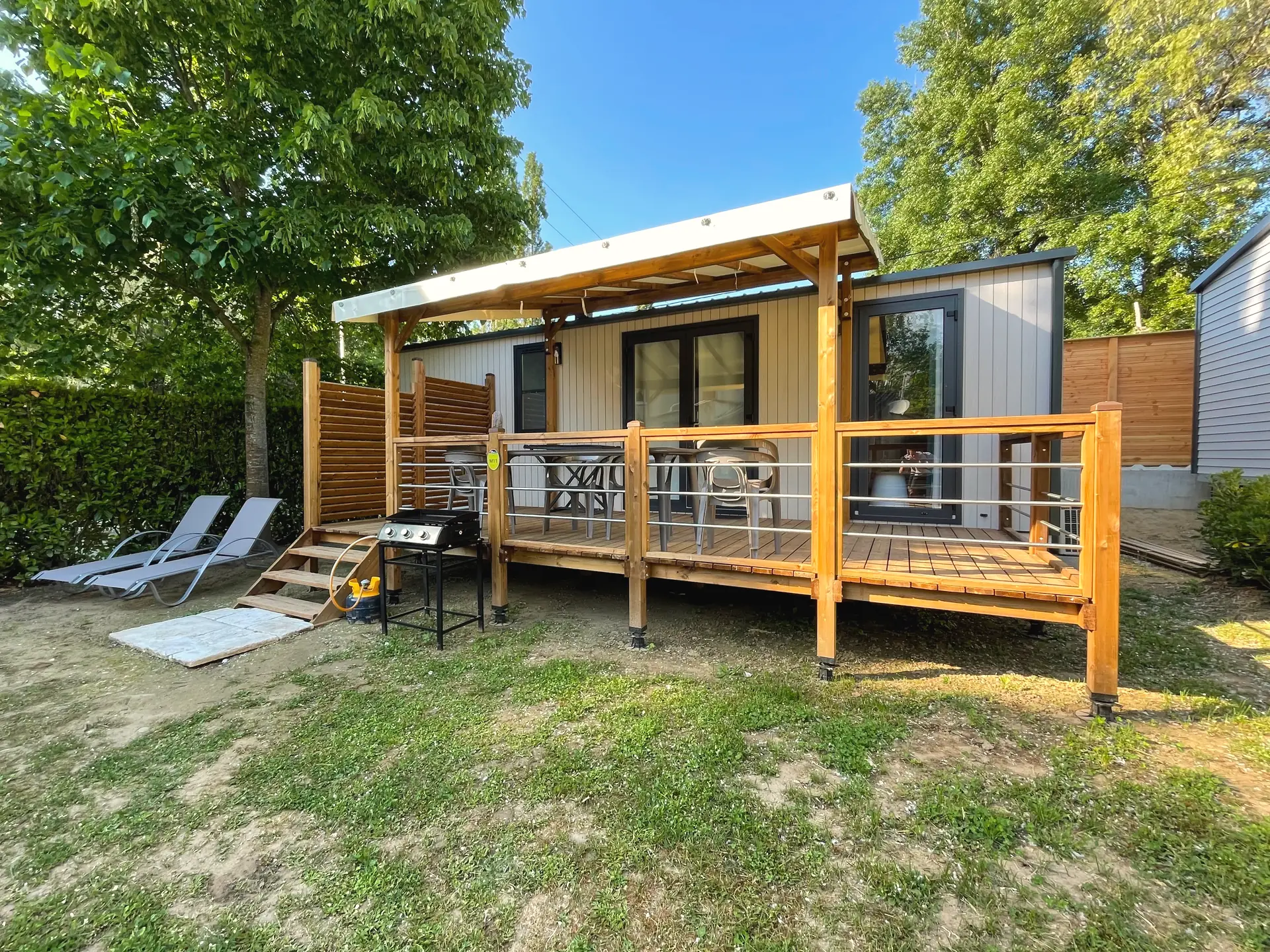 Ein Premium-Mobilheim mit Flachdach, seine Holzterrasse teilweise unter einer Pergola, zwei Sonnenliegen und eine Plancha auf dem Rasen
