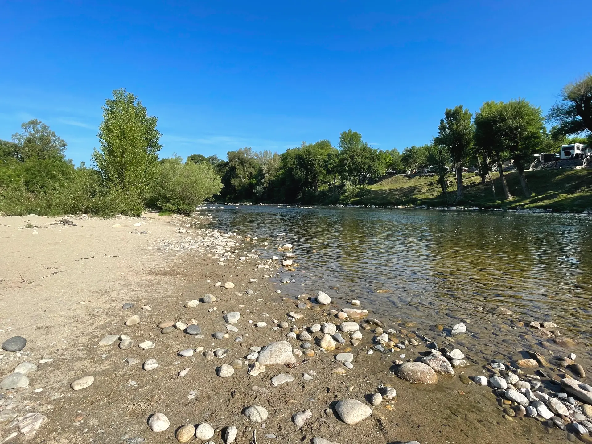 Plage de sable et de galets au bord de l'Ardèche, avec les emplacements du camping sous les arbres en arrière-plan