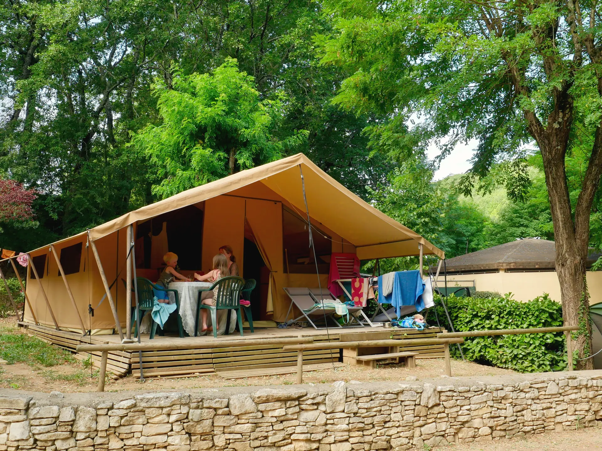 Tente Lodge et terrasse couverte avec une famille attablée, derrière un muret en pierres et sous les grands arbres